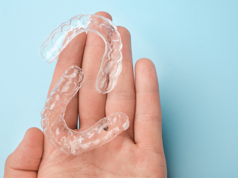 The image displays a hand holding two transparent dental aligners, molded to fit over the teeth for orthodontic treatment. The light blue background contrasts with the clear aligners, emphasizing their shape and function.