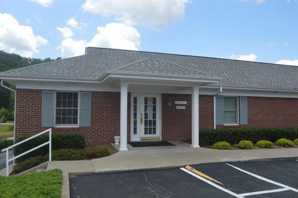 The image depicts a single-story brick building with a white columned porch, a glass-paneled front door, and two windows with light blue shutters. The surroundings include neatly trimmed bushes, a small lawn, and a paved parking area with marked spaces under a partly cloudy sky.